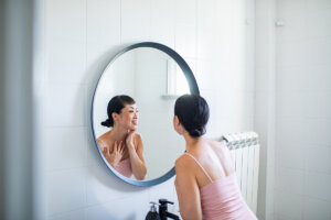 A woman smiling at her reflection in a circular mirror while touching her neck, promoting confidence and self-care in a minimalistic bathroom setting.