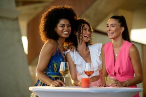 three women in dresses standing around table with cocktails laughing