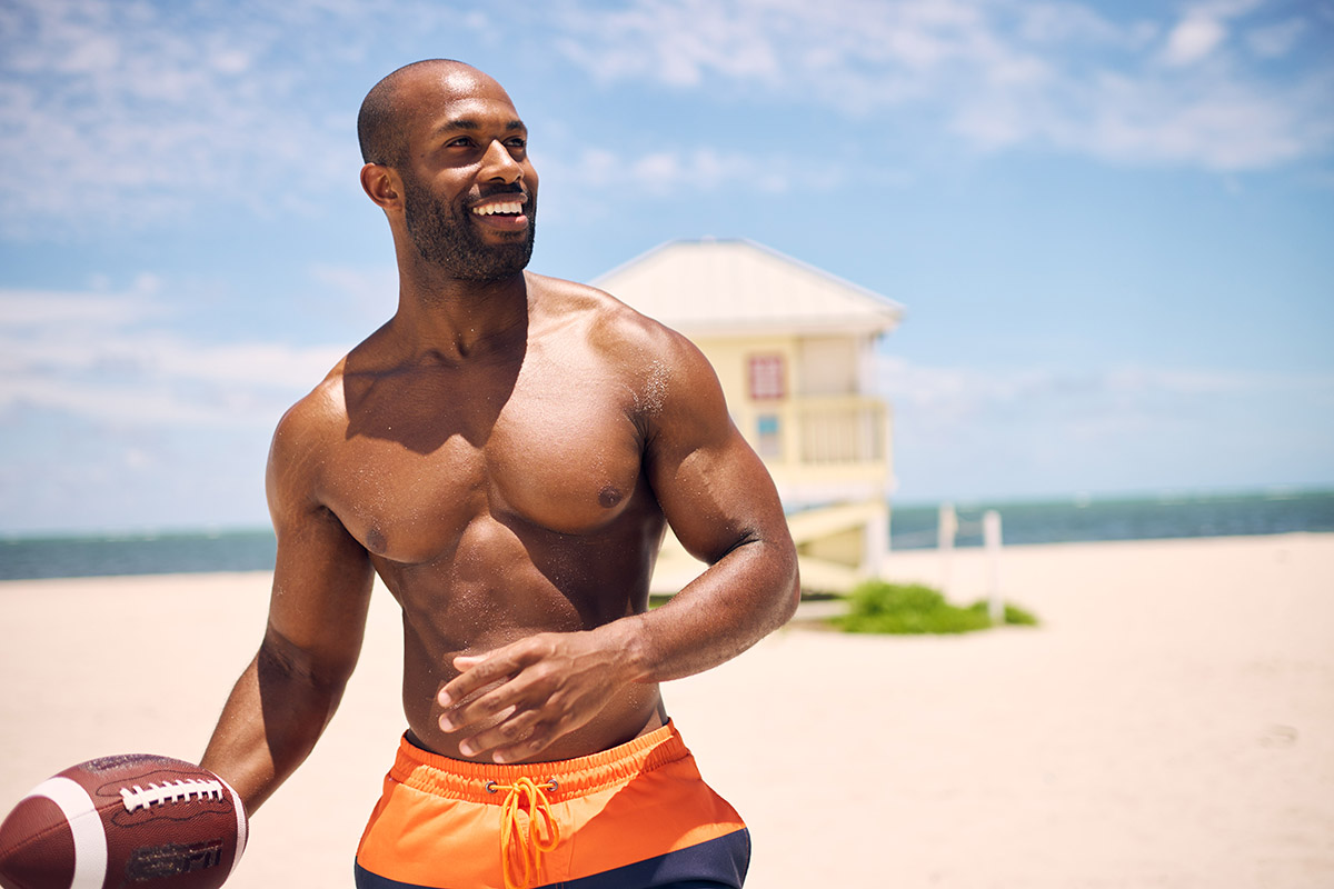 Fit man playing football on the beach, smiling under a bright blue sky.