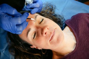 A woman lying on a salon table getting her brows laminated by a technician wearing blue latex gloves.