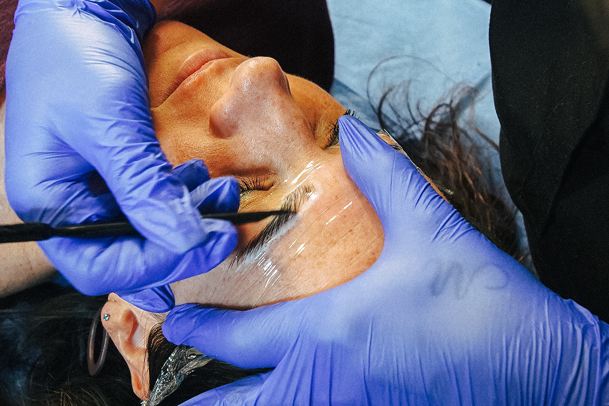 Close-up of a woman getting her brows laminated by a technician wearing blue latex gloves.