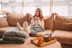 Woman sitting comfortably on a cozy beige sofa, holding a warm cup of tea, enjoying a relaxing moment indoors with folded sweaters and a candle nearby.