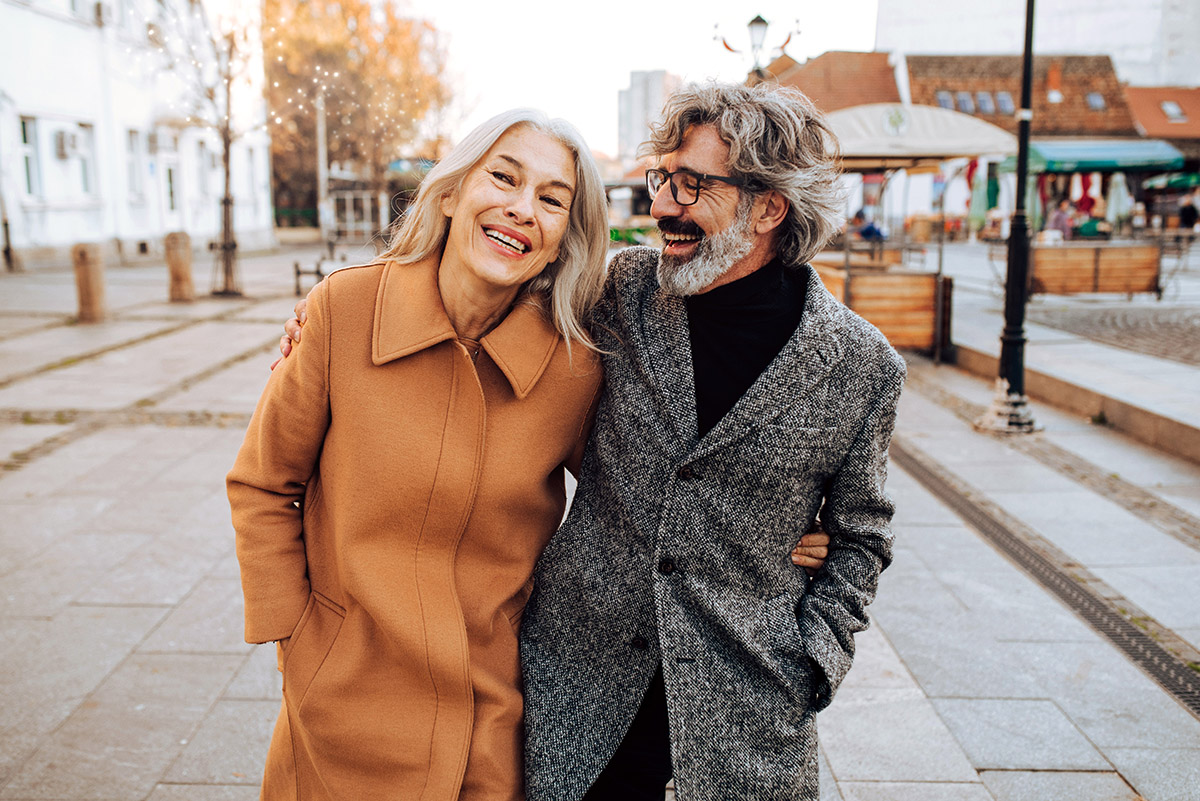 Joyful mature couple walking arm-in-arm outside, dressed in warm fall coats, enjoying a day out together with big smiles.