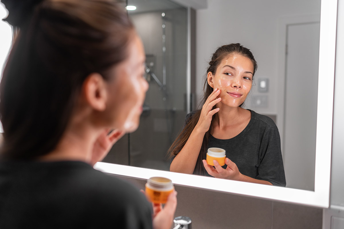 Woman applying moisturizing face cream while looking into a well-lit mirror, focusing on skincare and maintaining healthy skin.