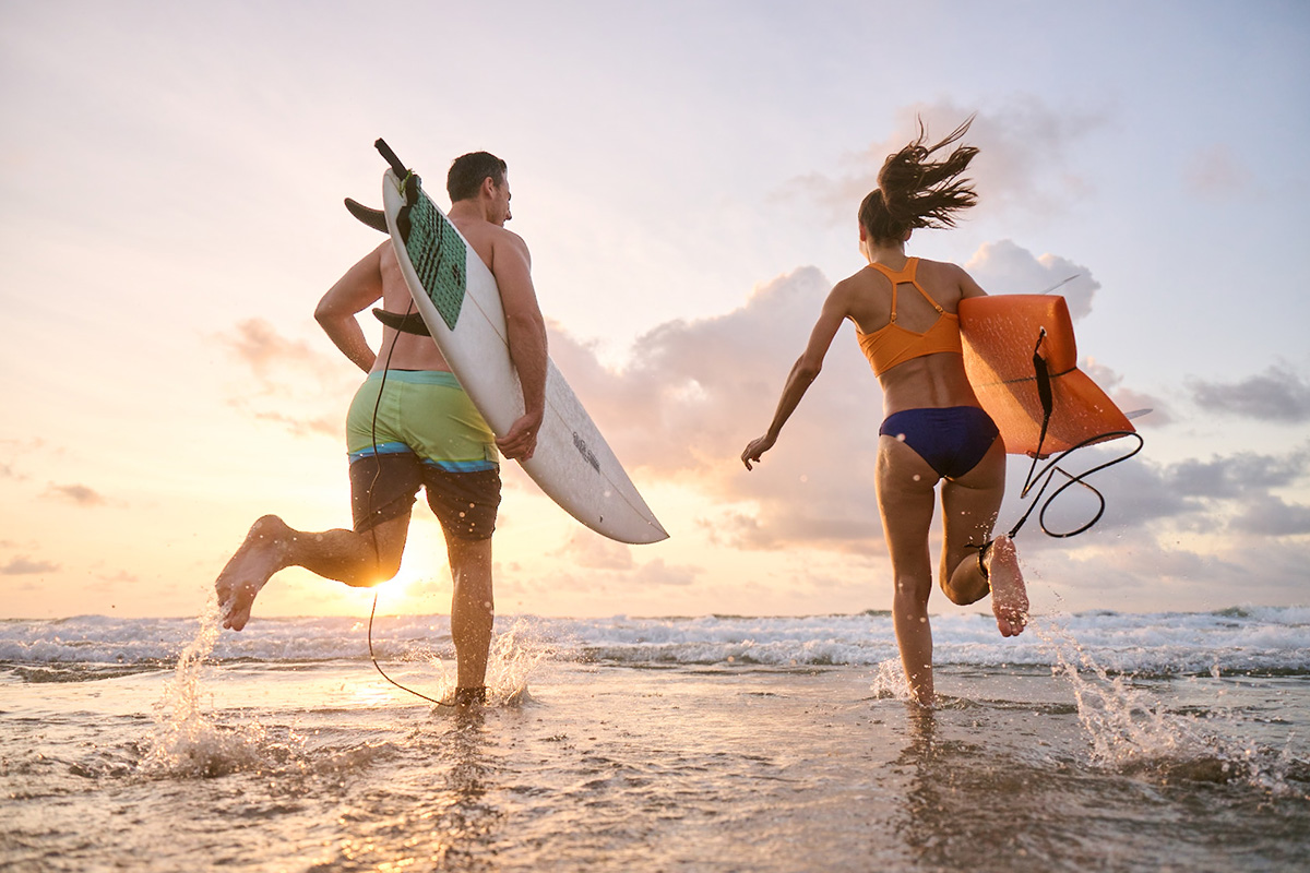 A man and woman running into the ocean with surfboards at sunset, highlighting the benefits of smooth, hair-free skin for an active lifestyle after waxing.