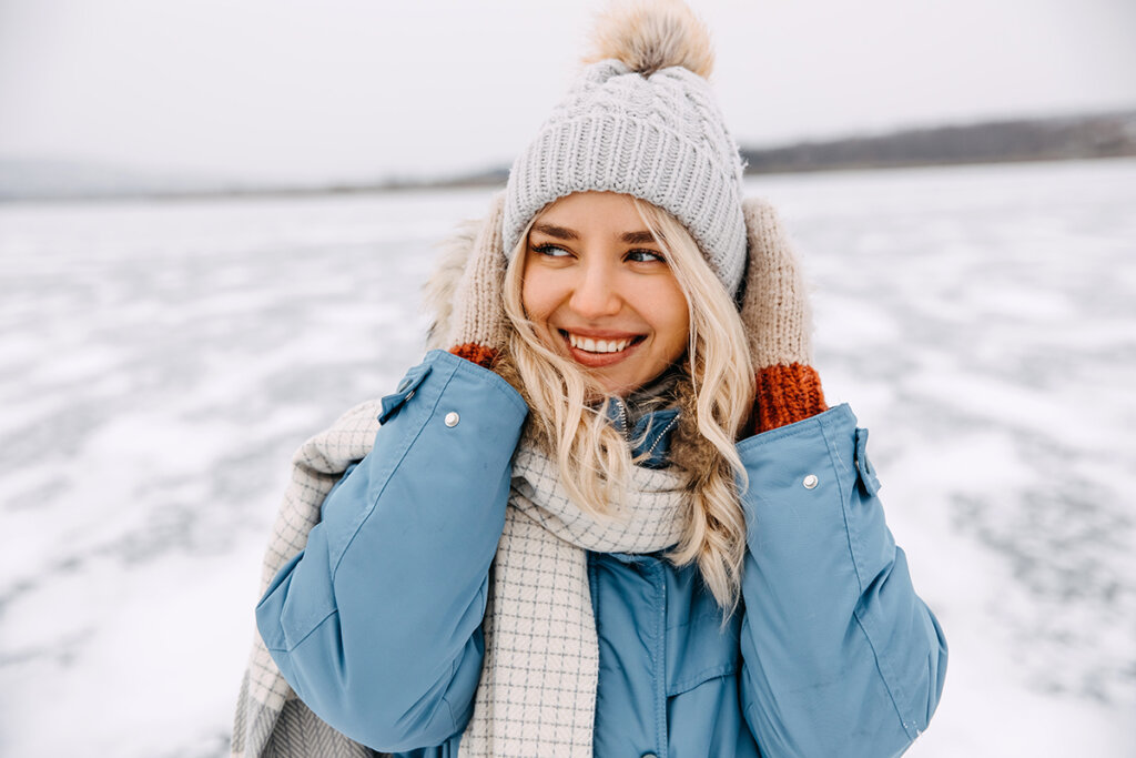 A smiling woman in a blue jacket, knit hat, and gloves enjoying winter outdoors, showcasing healthy, hydrated skin despite the cold weather.