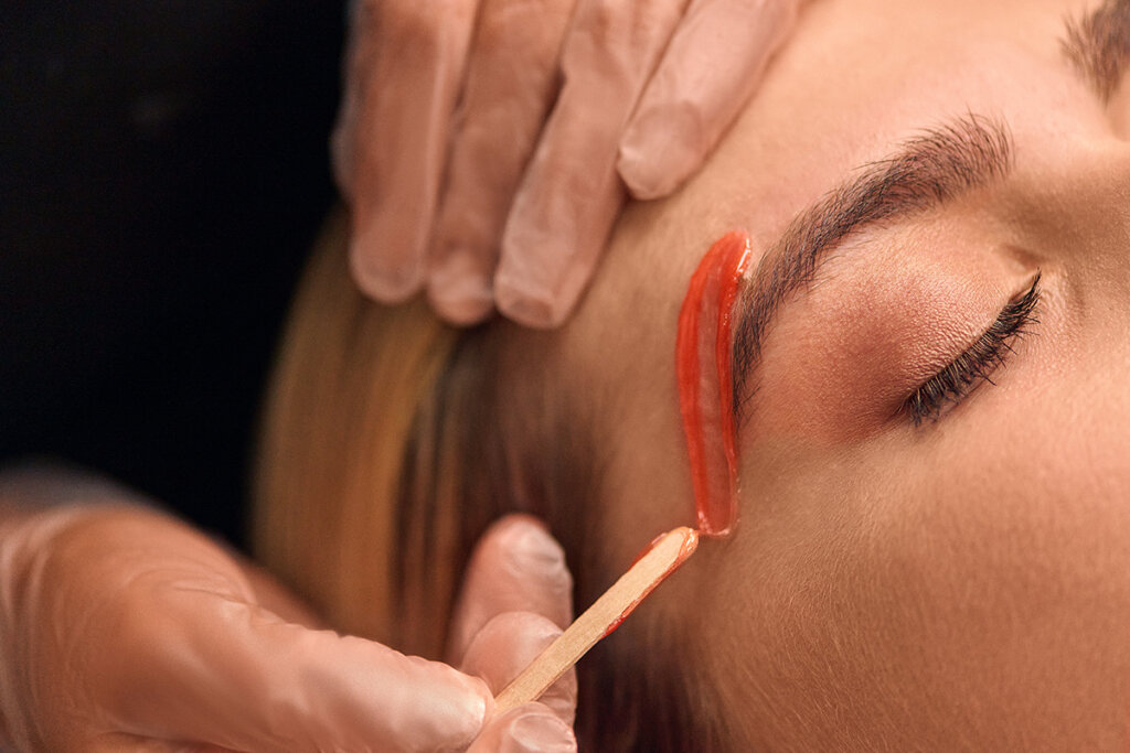 A woman is getting her brows waxed. A technician’s hands are shown applying red wax right above the brow.