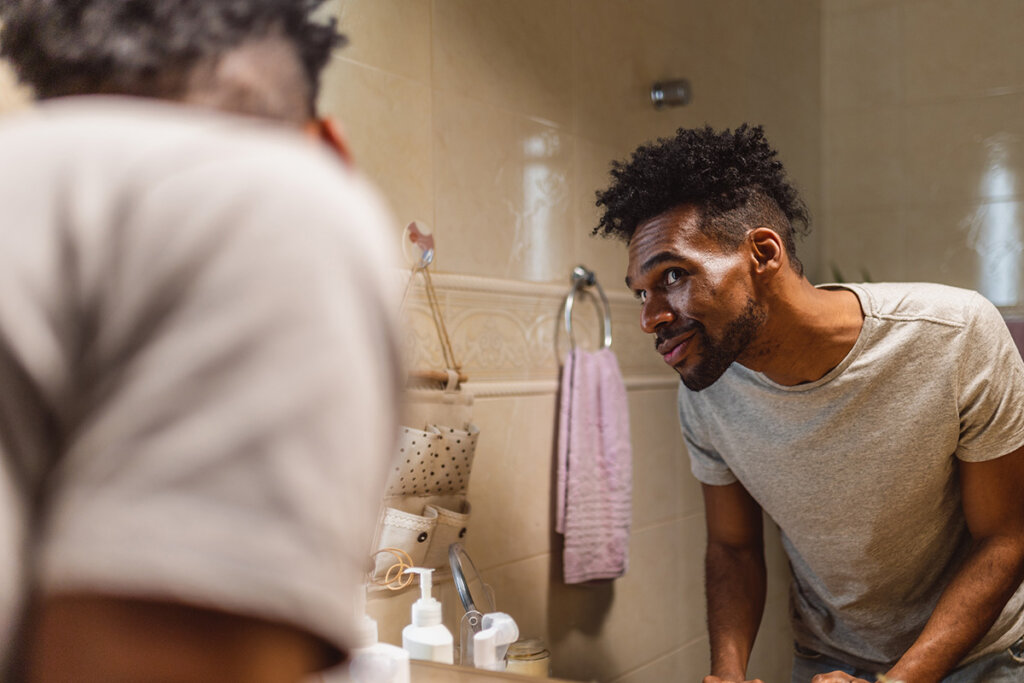 A man looks at himself in the mirror in a bathroom, while smiling and checking out his beard.